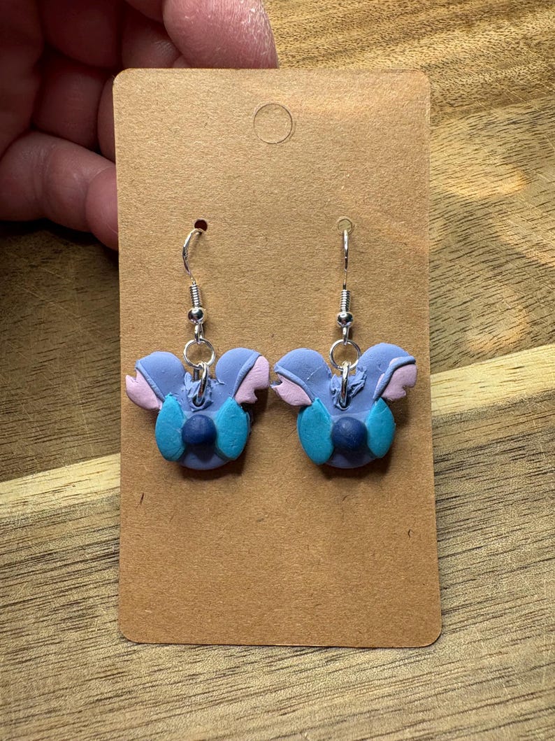 May include: A pair of handmade earrings featuring a blue, pink, and teal character design. The earrings are attached to silver-colored fish hook ear wires and are displayed on a brown card.