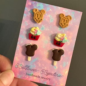 May include: A card with six handmade earrings. The earrings are shaped like Mickey Mouse, cupcakes, and ice cream bars. The card has a pink and purple background with the text "Bellflower Byouterie handmade jewelry."