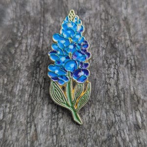 Könnte beinhalten: Eine blaue Lupinenblumenbrosche mit goldfarbener Umrandung. Die Brosche zeigt Blau- und Lilatöne mit grünen Blättern und einem gelben Stiel. Die Blume ist ein detailliertes Emaille-Design.