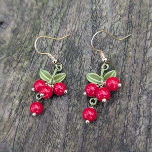 Pode incluir: Um par de brincos de ouro pendurados com esmalte vermelho e verde. Os brincos apresentam um cacho de bagas vermelhas com folhas verdes.