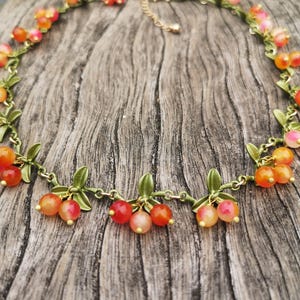 Op de afbeelding: Een ketting met een ontwerp van oranje en rode kralen fruitclusters met groene bladeren. De ketting heeft een goudkleurige ketting en sluiting. De kralen zijn een mix van oranje, rood en roze tinten, wat een levendige en naturalistische uitstraling creëert.