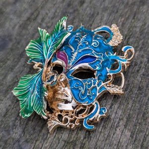 Op de afbeelding: Een goudkleurig masker voor een maskerade met blauwe en groene emaille verf. Het masker heeft ingewikkelde details en een grillig ontwerp.