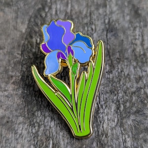 Op de afbeelding: Een goudkleurige metalen speld in de vorm van een irisfleur. De bloem heeft blauwe en paarse bloemblaadjes met groene bladeren. De speld is geplaatst op een verweerd houten oppervlak.