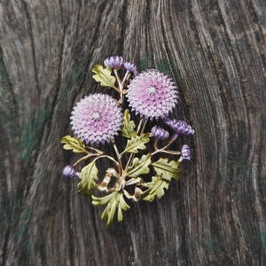 Könnte beinhalten: Eine Metallbrosche in Form eines Blumenstraußes mit zwei großen, runden, hellvioletten Blüten mit texturierten Mitten, umgeben von mehreren kleineren violetten Knospen und detaillierten grünen Blättern, verbunden durch dünne Stiele und eine kleine Schleife.