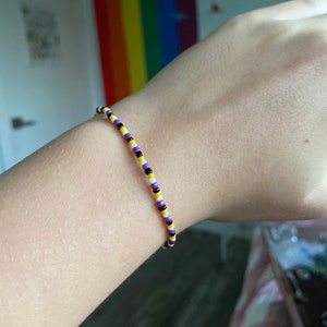 May include: A purple, yellow and black beaded bracelet on a person's wrist.