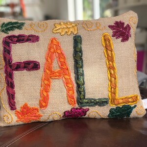 May include: Rectangular burlap pillow with the word "FALL" embroidered in colorful yarn. The letters are in shades of purple, orange, green, and yellow. Decorative leaves are embroidered around the letters.