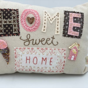 May include: A beige pillow with the word "HOME" spelled out in brown and pink fabric letters. The letters are surrounded by pink and brown fabric accents. The words "Sweet Home" are embroidered in brown thread on the pillow.