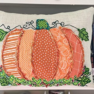May include: A beige pillow with an embroidered pumpkin design. The pumpkin is made of various orange fabrics with different patterns, including stripes, polka dots, and a floral print. The pumpkin is surrounded by green leaves.