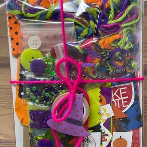 May include: A clear plastic bag filled with Halloween craft supplies, including colorful yarn, fabric scraps, buttons, beads, and a witch hat cutout. The bag is tied with a pink ribbon.