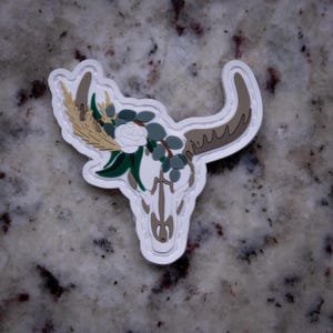 May include: A white, tan, and brown bull skull patch with floral accents. The patch features a white rose, green leaves, and tan wheat stalks. The skull has brown horns and a white outline.