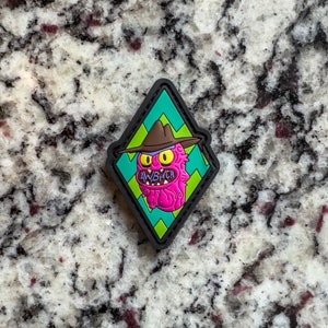 May include: A black diamond-shaped patch with a green and yellow chevron pattern. The patch features a pink cartoon character with a brown hat and the text "AWB/CH".