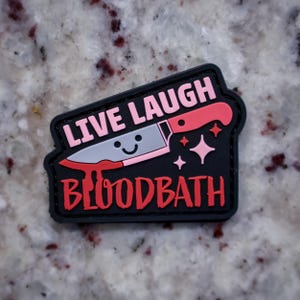 Puede incluir: Un parche negro con las palabras "LIVE LAUGH BLOODBATH" en rosa y rojo. Un cuchillo sonriente con sangre goteando de la hoja está en el centro. Estrellas rosas están a la derecha del cuchillo.