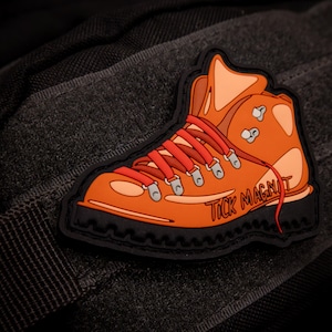 Könnte beinhalten: Orangener Wanderstiefel-Aufnäher mit roten Schnürsenkeln und den Worten "TICK MAGNET" in Weiß. Der Aufnäher hat einen schwarzen Rand und ist vor einem dunkelgrauen, strukturierten Hintergrund platziert. Das Stiefeldesign ist im Cartoon-Stil gehalten.