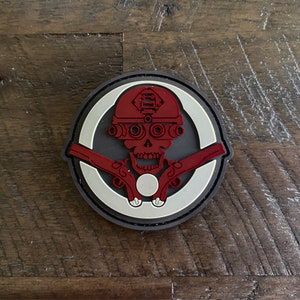 Könnte beinhalten: Ein roter und schwarzer Schädel mit Helm und Schutzbrille hält zwei Pistolen. Der Schädel ist von einem weißen Kreis mit schwarzem Rand umgeben. Der Hintergrund ist eine braune Holzoberfläche.