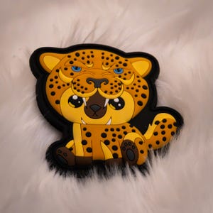 May include: A cartoon-style leopard patch with a black border. The leopard is yellow and orange with black spots, blue eyes, and a brown mouth. The patch is sitting on a white, fluffy surface.