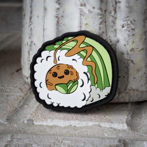 Può includere: Una toppa colorata a tema sushi con un design a cartoni animati. La toppa presenta un personaggio marrone sorridente all'interno di un rotolo di sushi con riso bianco, alghe verdi e un filo di salsa marrone. La toppa ha un bordo nero.