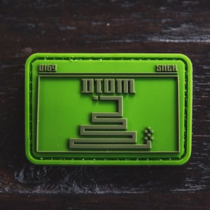 Puede incluir: Parche bordado verde y marrón con el texto "DIOM" y un diseño de serpiente pixelado. El parche tiene el texto "0169" y "5N6H" en las esquinas superiores.