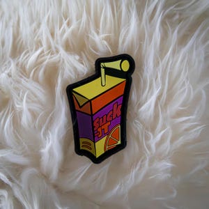 Può includere: Una scatola di succo viola, arancione e gialla con una cannuccia e il testo "Suck It" stampato sul davanti. La scatola di succo è su una superficie bianca e soffice.