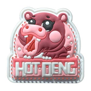 May include: A pink and maroon hippo-shaped charm with the text "HOT DENG" in white letters. The charm has a white border with a stitched design. The hippo has a wide open mouth.