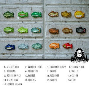 May include: A selection of embroidered fish patches in various colours and designs, including species such as Atlantic Cod and Rainbow Trout. The patches are displayed on a textured, light grey surface. Each patch is approximately 5 cm in length.