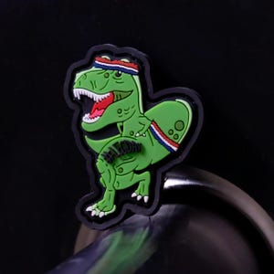 May include: A green dinosaur-shaped charm with a red mouth, white teeth, and a red, white, and blue headband. The charm has the text "#LEG DAY" and is outlined in black.