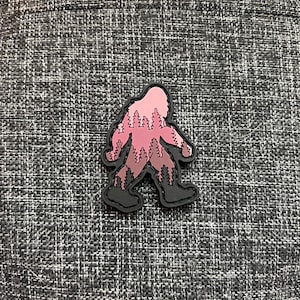 Puede incluir: Un parche negro y rosa con la silueta de Bigfoot con un patrón de bosque en su interior. El parche está hecho de un material duradero y es perfecto para agregar un toque de fantasía a tus pertenencias.