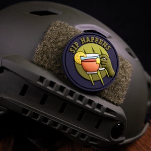 May include: A circular morale patch attached to an olive drab tactical helmet. The patch features a dark blue border with the text SIP HAPPENS in yellow, surrounding a green background with a tea cup, a lemon slice, and a hand holding the cup.