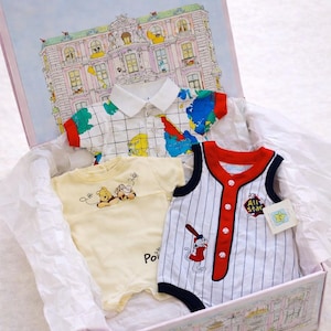 May include: A gift box containing three baby outfits. One is a white polo shirt with a colorful map design. Another is a yellow onesie with a Winnie the Pooh graphic. The third is a baseball-themed romper with red accents.
