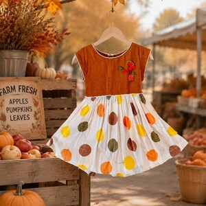 May include: A dress with a solid rust-colored bodice featuring a small embroidered fruit cluster on the chest and a white gathered skirt patterned with large, scattered polka dots in shades of yellow, orange, brown, and olive green.