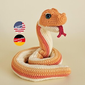 Op de afbeelding: Een gehaakt slangenfiguur met een opgerold lichaam in oranje, crème en lichtbruine garentexturen. De slang heeft een opgeheven kop, donkergroene ogen en een uitstekende rode gespleten tong. Twee ronde iconen tonen English PDF en Deutsche PDF.