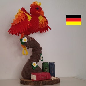Könnte beinhalten: Ein gehäkelter Phönix mit roten, orangen und gelben Federn, der auf einem braunen gehäkelten Baumstamm sitzt. Der Vogel hält ein Buch in seinen Krallen. Der Baumstamm ist mit weißen gehäkelten Blumen verziert. Auf einem Holzsockel liegen drei Bücher übereinander.