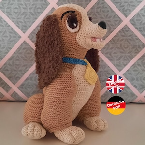 Könnte beinhalten: Gehäkeltes Plüschtier von Lady, dem Hund aus Disneys Lady und der Tramp. Das Spielzeug ist braun und weiß mit einem blauen Halsband und einem gelben Anhänger. Das Spielzeug sitzt auf einer grauen und rosa gemusterten Oberfläche. Das Bild enthält auch Text in Englisch und Deutsch für das PDF-Muster.