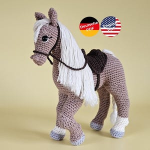 Peut inclure: Un cheval en crochet aux tons marron, blanc et gris. Le cheval a une crinière et une queue blanches, une selle marron et un bridon. Le jouet est fait à la main et se tient sur quatre pattes. L'image comprend deux graphiques circulaires avec le texte "Deutsche PDF" et "English PDF".