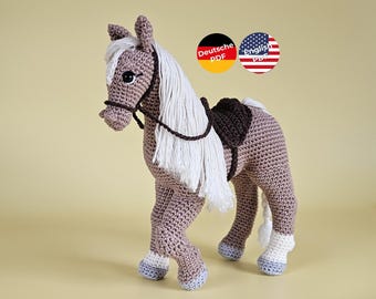May include: A crocheted horse toy in shades of brown, white, and gray. The horse has a white mane and tail, a brown saddle, and a bridle. The toy is handmade and stands on four legs. The image includes two circular graphics with the text "Deutsche PDF" and "English PDF".