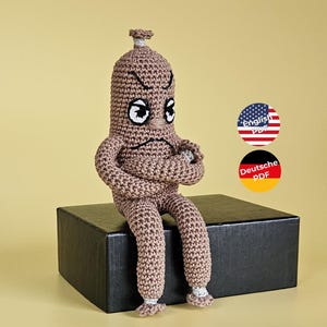 Könnte beinhalten: Eine handgefertigte, gehäkelte Figur in Brauntönen mit einem wütenden Gesichtsausdruck. Die Figur hat verschränkte Arme und sitzt auf einer schwarzen Box. Die Figur hat weiße Augen und trägt weiße Socken. Der Hintergrund ist hellgelb.