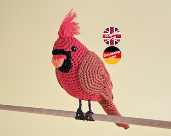 May include: Crocheted cardinal bird toy in red and brown, perched on a wooden dowel. The toy has black eyes and a black beak with an orange tip. The image includes text in English and German, indicating that the pattern is available in both languages.