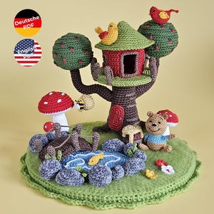 Könnte beinhalten: Eine skurrile, gehäkelte Baumhausszene mit einem roten und grünen Haus, einem Teich und einer Bärenfigur. Die Szene umfasst einen Baum mit grünen, gehäkelten Kugeln, einen Teich mit blauem Wasser und einen Bären mit blauem Hemd.