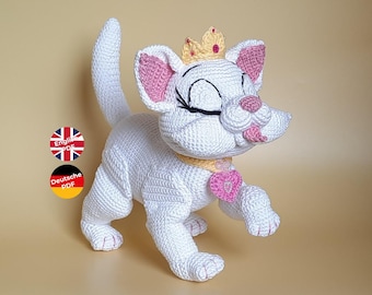 May include: White crocheted cat with pink ears and a yellow crown. The cat is wearing a yellow collar with a pink heart charm. The cat is standing on a light brown surface.