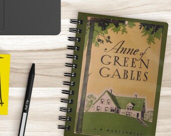 May include: A spiral-bound notebook with the title "Anne of Green Gables" and a green and tan cover, next to a black pen and a yellow sticky note with doodles. A black laptop is in the background.
