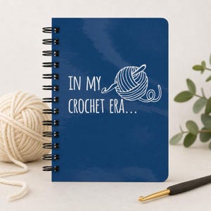 May include: A dark blue spiral-bound notebook featuring a white graphic of a ball of yarn with a crochet hook and the text "IN MY CROCHET ERA..." printed in white on the front cover. The notebook is positioned next to a ball of yarn.