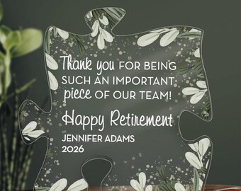 May include: Clear acrylic puzzle piece with white text: "Thank you for being such an important piece of our team! Happy Retirement Jennifer Adams 2026." The piece has a floral design with white leaves and green accents.
