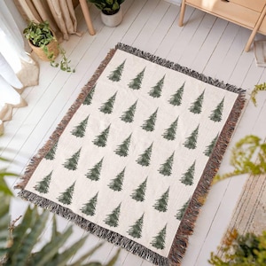 May include: A woven throw blanket with a white background and a repeating pattern of dark green stylized pine trees. The blanket has fringe around the edges.
