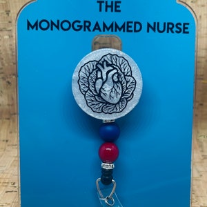 May include: A silver heart with a black anatomical design inside a leafy wreath, attached to a blue and red beaded retractable badge holder with the text "THE MONOGRAMMED NURSE".
