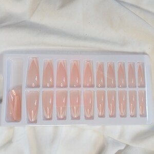 Puede incluir: Un juego de 20 uñas postizas de color nude en forma de ataúd con punta blanca. Las uñas están dispuestas en una bandeja de plástico blanca.