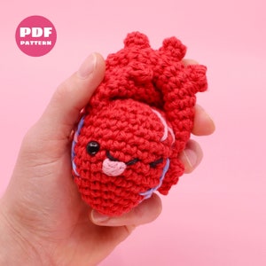 May include: A crocheted red heart with a cute face, blue and pink details, and a winking eye. The heart is being held in a hand against a pink background.  PDF PATTERN.