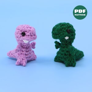 Puede incluir: Dos juguetes de dinosaurio de crochet, uno rosa y otro verde, con ojos negros y dientes blancos. Los juguetes están sobre un fondo azul claro. PATRÓN PDF.