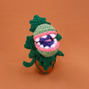 May include: A handmade crochet plant with a large, open mouth. The plant features a green head with pink and white accents, and a purple interior. The plant is in a brown pot, with green leaves surrounding it. The background is a solid orange color.