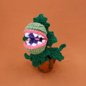 May include: A handmade crochet plant with a large, open mouth. The plant features a green head with white teeth, a pink and green mouth, and a purple interior. The plant is in a brown pot with green leaves and is set against an orange background.