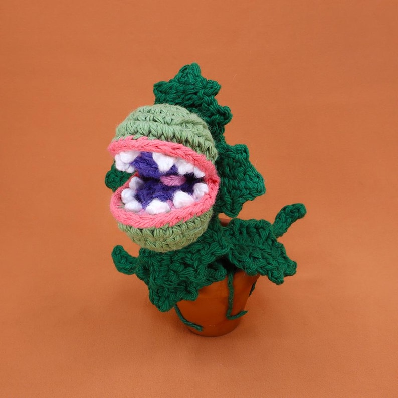 May include: A handmade crochet plant with a large, open mouth. The plant features a green head with white teeth, a pink and green mouth, and a purple interior. The plant is in a brown pot with green leaves and is set against an orange background.