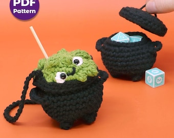May include: Crocheted black cauldron with a green monster face, a wooden stick, and a black handle. A second black cauldron with a lid is open, revealing blue dice. A single die is also visible. The image includes a purple circle with the text "PDF Pattern".